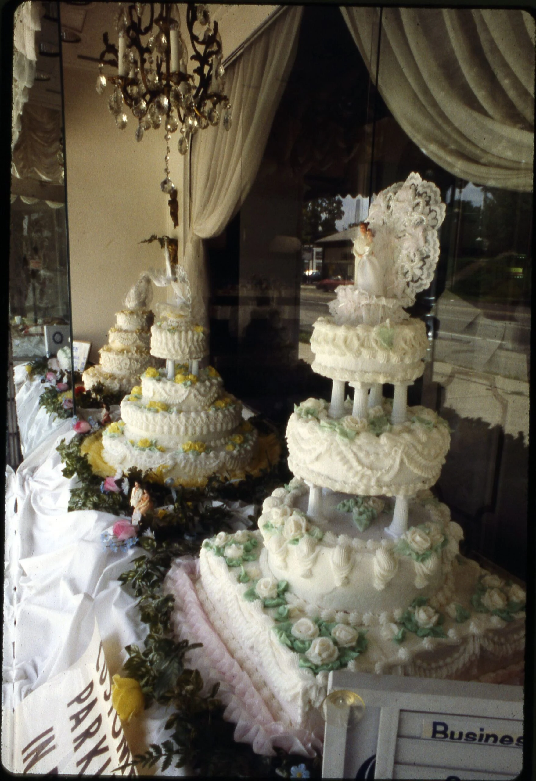 Vitrine de boulangerie vintage avec trois gâteaux de mariage à plusieurs niveaux sur des piliers blancs, décorés de crème au beurre à volants, de guirlandes et de décorations en dentelle, sous un lustre