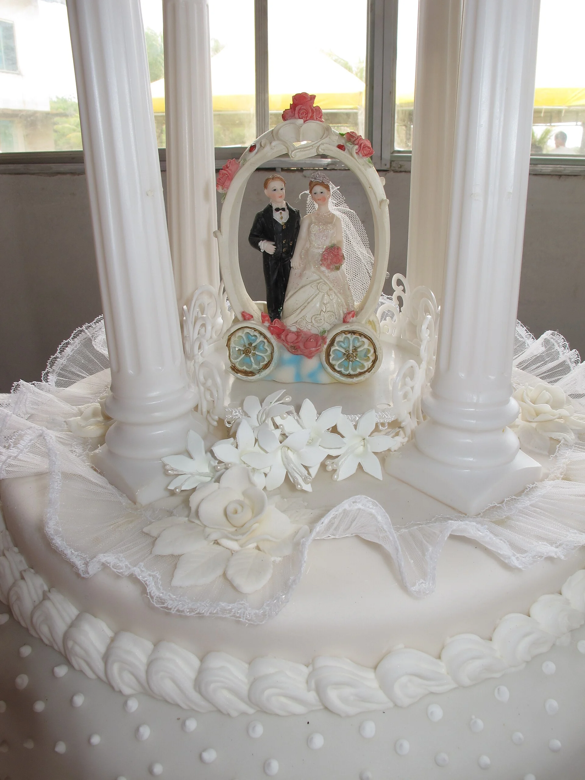 Décoration de gâteau de mariage représentant les mariés, encadrée à l'intérieur d'un arc ovale blanc, placée entre des colonnes blanches sur un gâteau à volants avec des détails en dentelle et en perles.