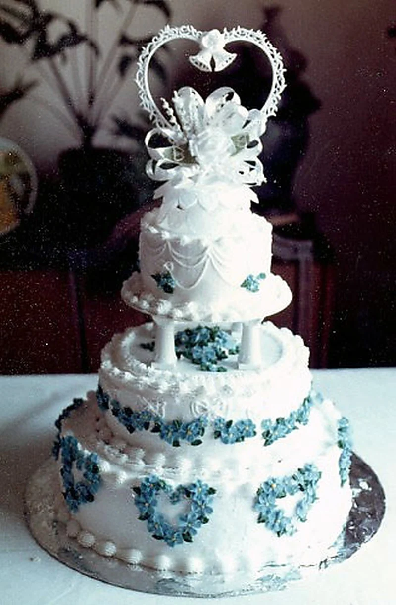 Gâteau de mariage à trois niveaux de style années 1980 avec glaçage blanc, bordures passepoilées bleues et accents de fleurs bleues, surmonté d'un cadre en forme de cœur