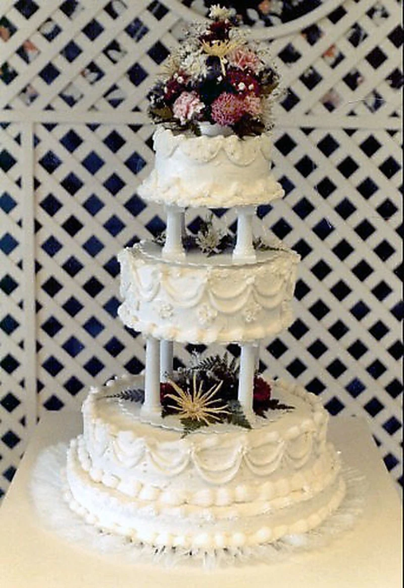 Gâteau de mariage à trois niveaux séparés par des piliers grecs blancs avec des bordures de coquillages et des guirlandes de glaçage en cascade, photographié sur un treillis blanc