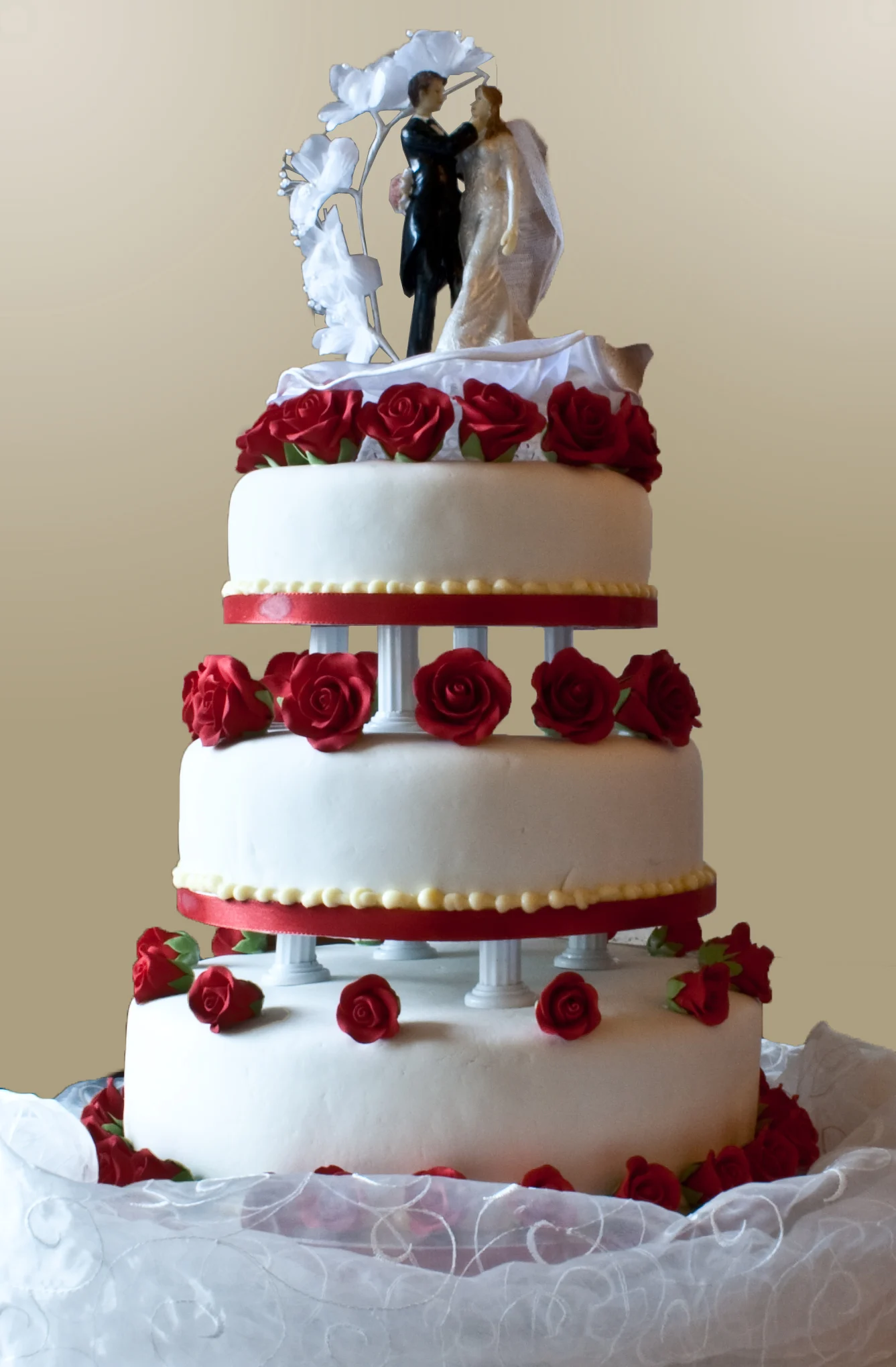 Gâteau de mariage à plusieurs niveaux sur de courts piliers avec des roses rouges entre les niveaux et une décoration pour les mariés sous une arche blanche