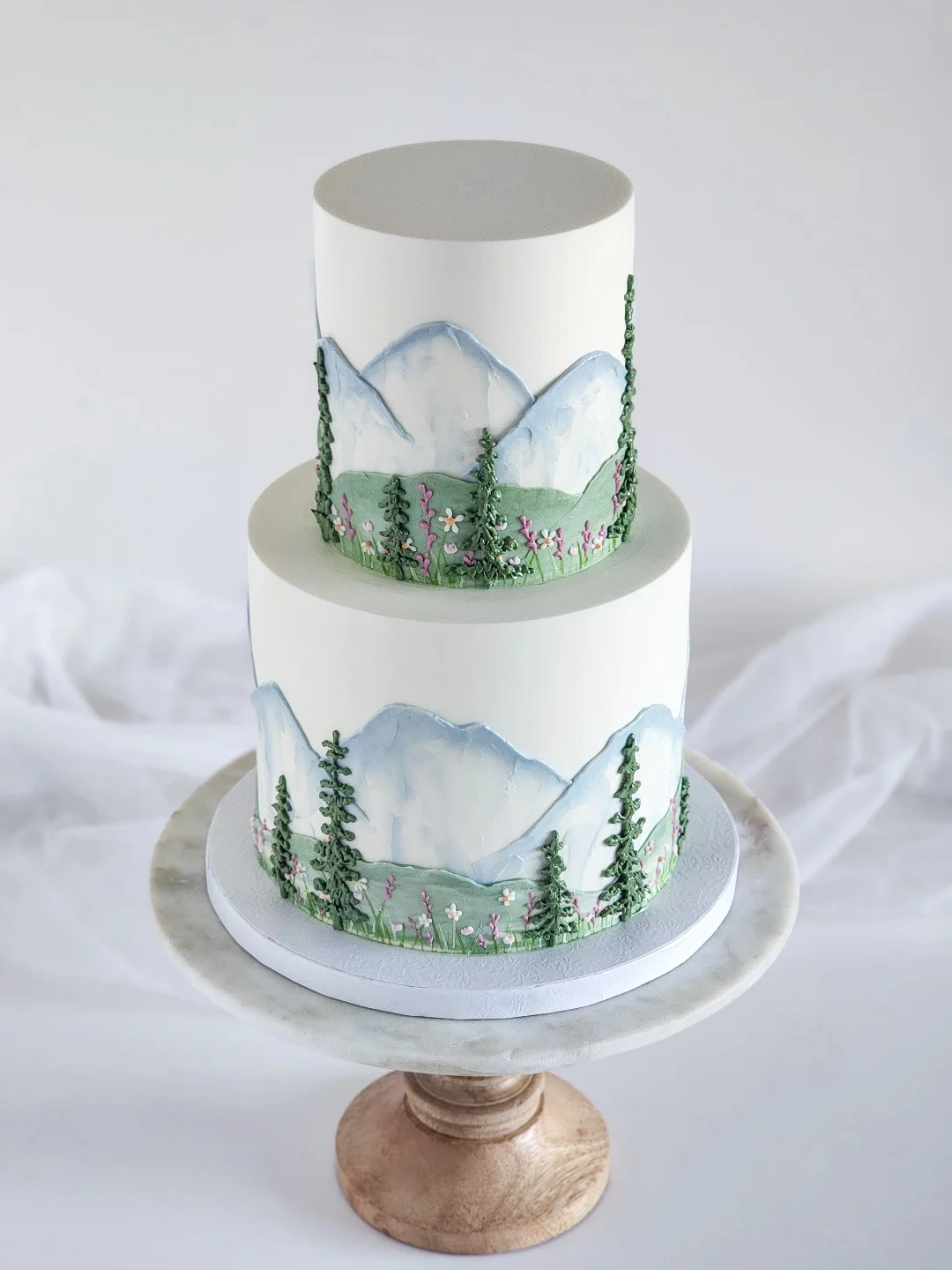 Gâteau de mariage blanc à deux niveaux peint avec des montagnes à l'aquarelle, des conifères et des fleurs sauvages