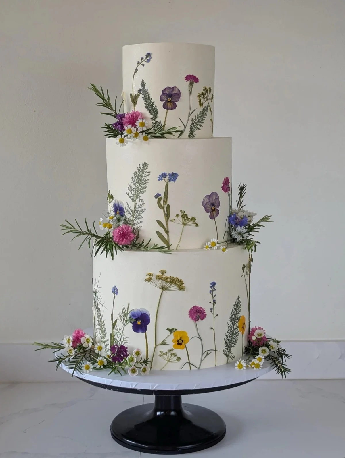 Gâteau de mariage blanc à trois niveaux décoré de fleurs sauvages comestibles pressées, notamment de pensées et de camomille